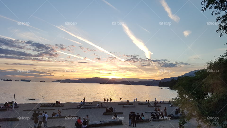 people sitting on logs on the beach overlooking English Bay in Vancouver at sunset. clear sky with streaks of cloud