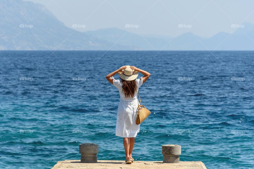 Rear view of young woman wearing white dress and sun hat standing on pier while admiring amazing sea