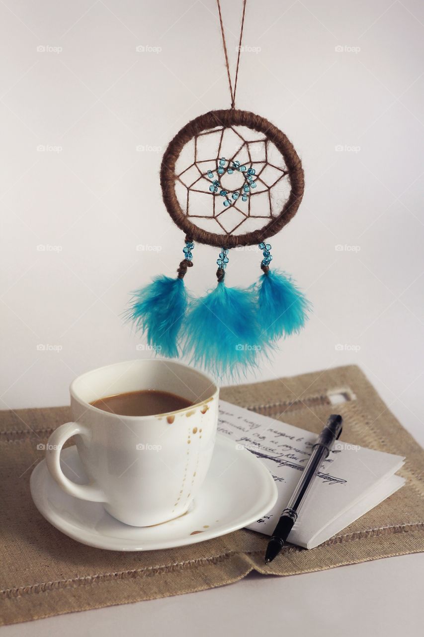 Still life of coffee in a white cup, dream catcher with blue beads and feathers and unfinished verses