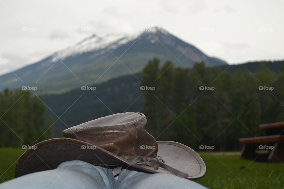 Snow capped Rocky Mountain in Canada in the background, person sitting viewing, only showing legs and leather cowboy hat foreground 