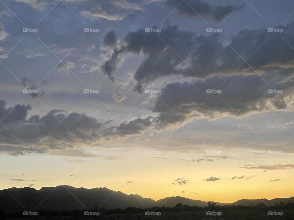 A bright yellow and orange sunset with several dark grey clouds above and mountains.
