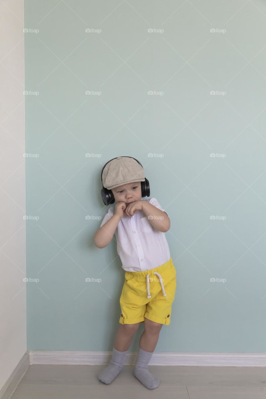 A little boy in yellow shorts, a white shirt and a cap listens to music in big, black headphones.  in retro style.