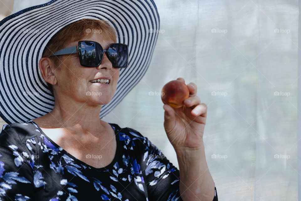 Portrait of one beautiful Caucasian elderly woman in sunglasses and straw hat with happy smile holding ripe peach in hand while sitting in wicker chair in garden against gauze curtain background on summer day, side view close up.