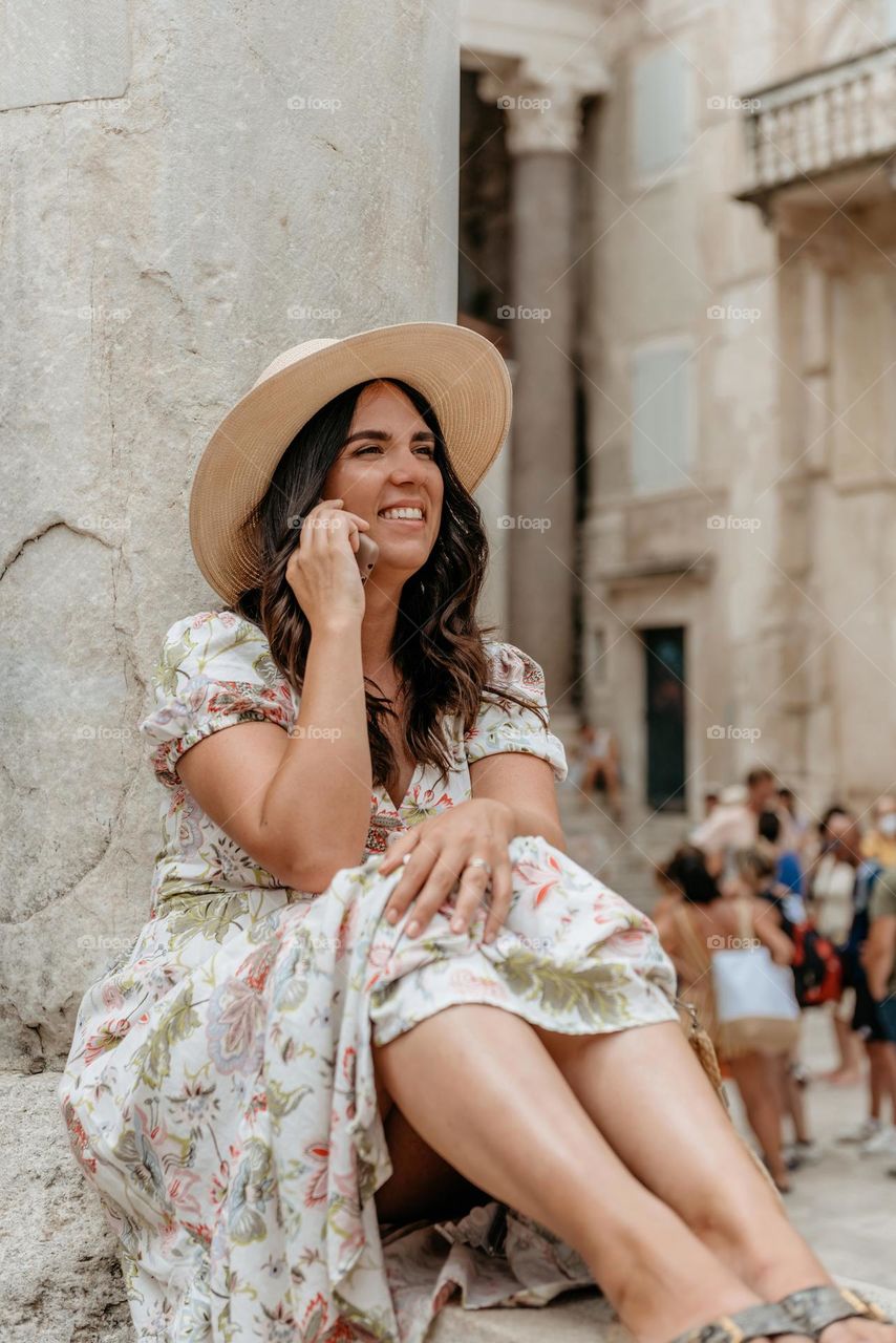 Happy young woman talking on mobile phone while sitting by column of ancient building in city