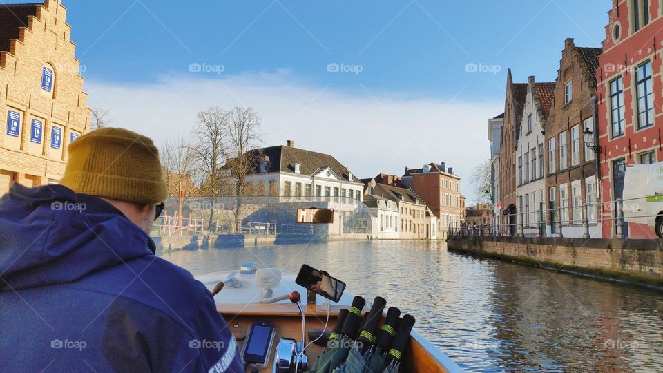 A caucasian man from the back in a mustard hat, blue jacket sits at the helm of a motor boat and floats forward along a canal with European-style houses of the sixteenth and nineteenth centuries to the city of Bruges in Belgium, vtd side close-up.