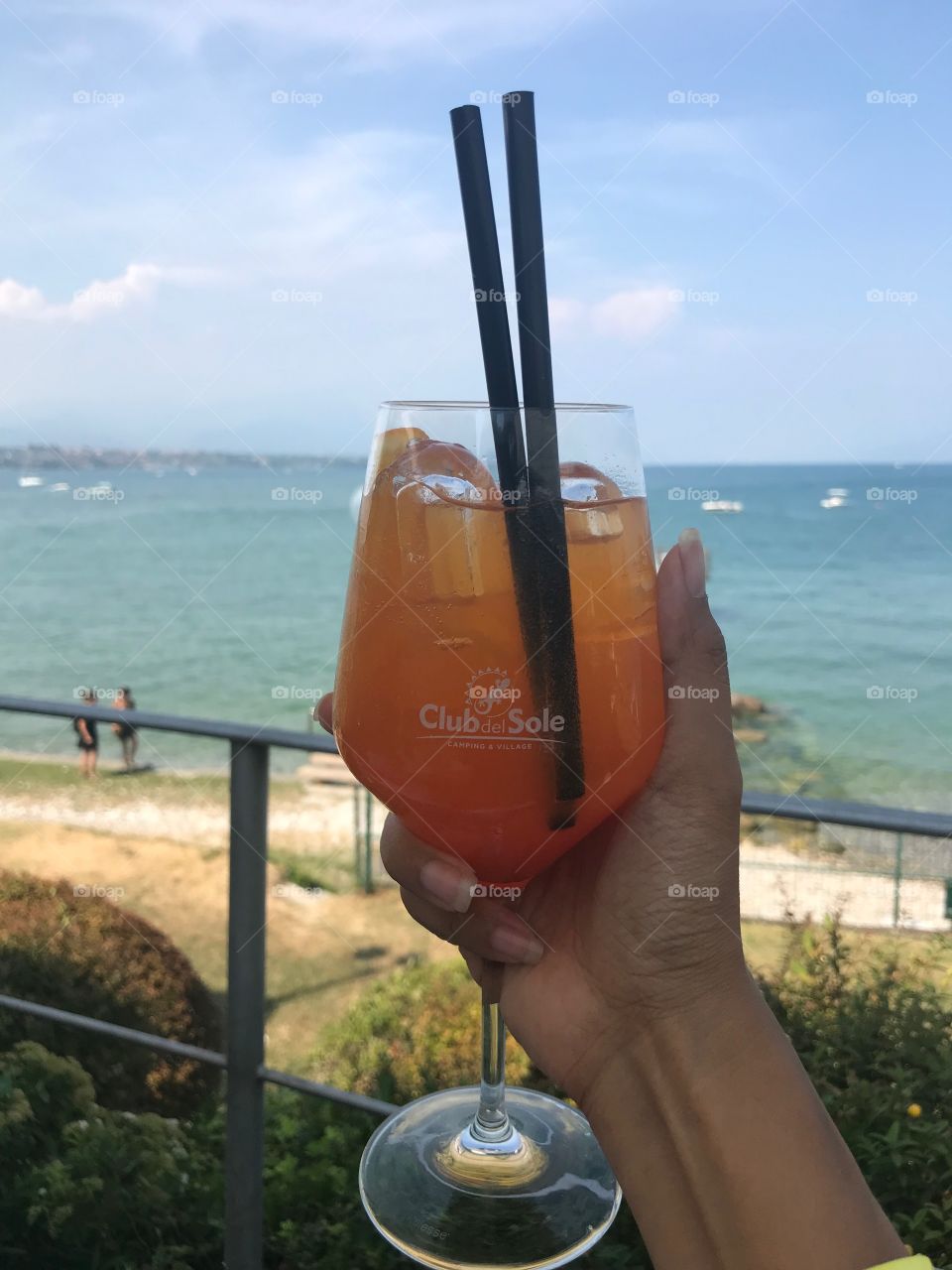 I holding a nice orange cocktail in my hand, Sitting above the restaurant overlooking the lake, There are many stones on the shore of the lake, the water has a very beautiful blue color. There is a couple walking, and a few canoes in the distance