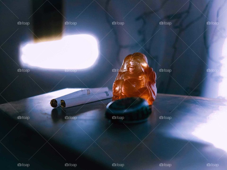 Little orange Buddha statue, with two cigarettes, beer bottle caps, with distinct lighting, detail shot.