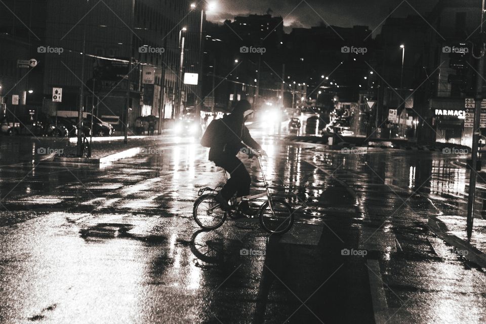 Black and white photo of a Man on bicycle crossing the street on zebra lines, on a rainy day in January of 2019, two cars are lighting the back of the subject, showing only a cihlouette of the bicycle and the man riding it.