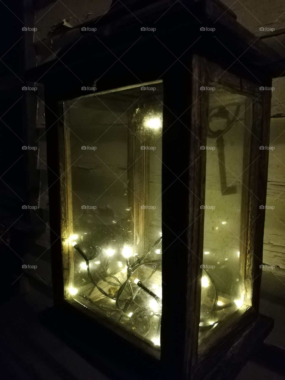 A square shape lantern hanging on the wall and having bright lights inside. An iron key hanging in the nail in the background.