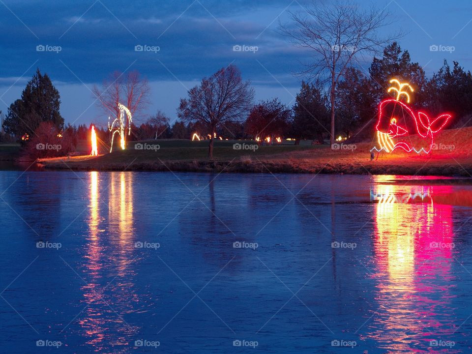 A whale display of Christmas lights reflecting on an iced over pond at dusk. 