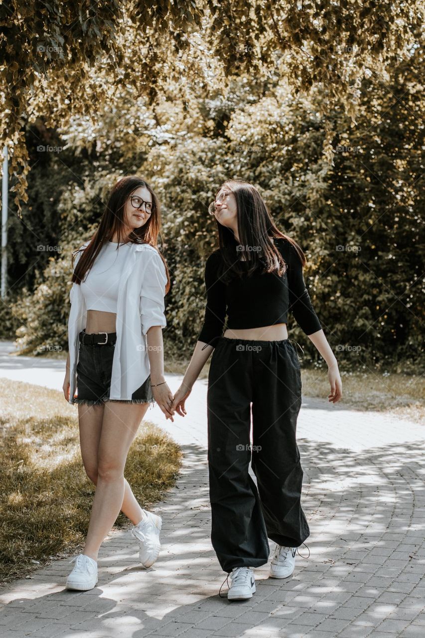 Portrait of two beautiful young caucasian teenage girls walking in the park on a summer day holding hands and gently looking into each other's eyes, close-up side view.