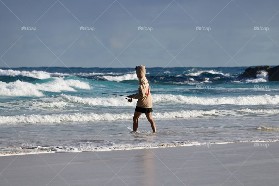 Person in hoodie and shorts fishing in wild water on remote beach coastline at Coffin Bay National Park in South Australia, crashing white water waves off reefs, turquoise sea blue sky, Australian wonders and outdoors 