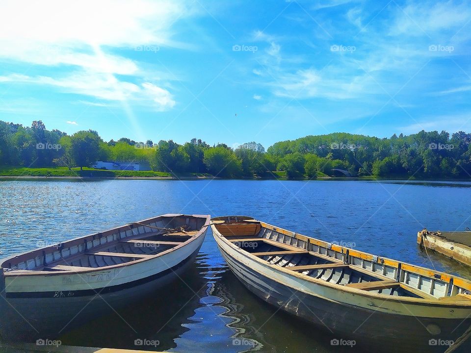 Two wooden boats are moored to the shore of the pond.  Blue water.  Blue sky with white clouds.  Green forest in the background