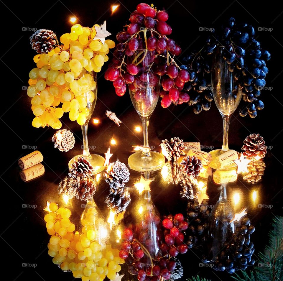 Three glasses with bunches of grapes in the form of hair stand on a mirrored surface. Yellow, red and black grapes. Around a luminous garland, pine cones and stars. Wine corks lie at the stems of the glasses. Reflection in the mirror