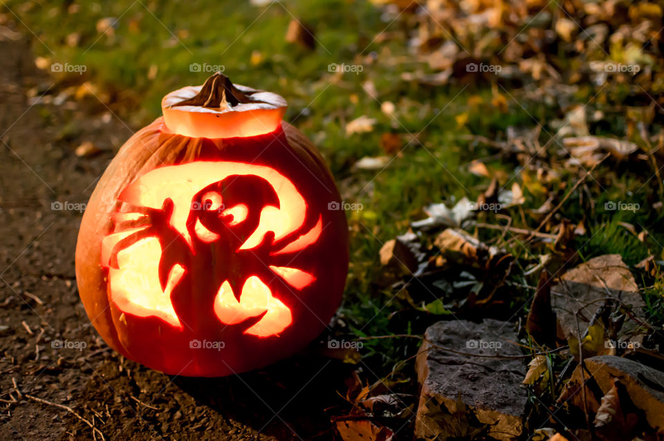 Beautiful carved pumpkin jack o’lantern outdoors in front yard to welcome trick or treaters with happy smiling ghost homemade creative pumpkin art background on grass with autumn leaves 
