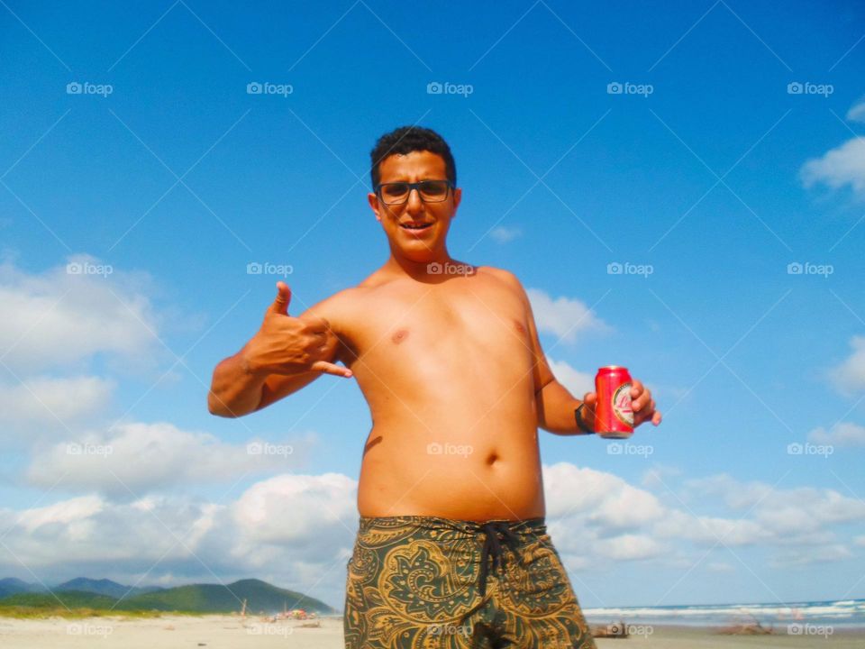 Man drinking a can of beer in the beach, with blue skies and clouds on the background.