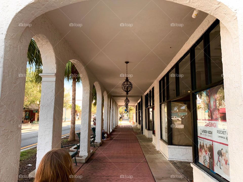 Facade walkway patio type architectural building with many arched doorways in perspective with chandelier in the middle and girl walking in tropical environment.