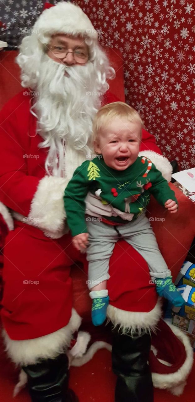 terrified of Santa , 1 year old baby sat on Santa's knee for first time , scared . Crying baby in Christmas jumper.