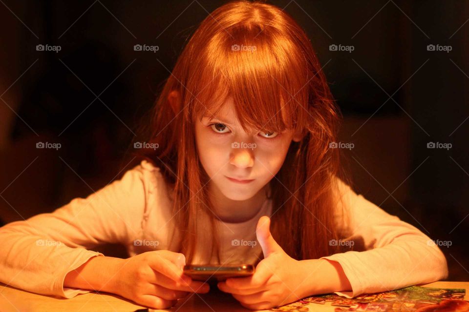 Little girl sits at a table with a gadget in her hands by the light of a table lamp