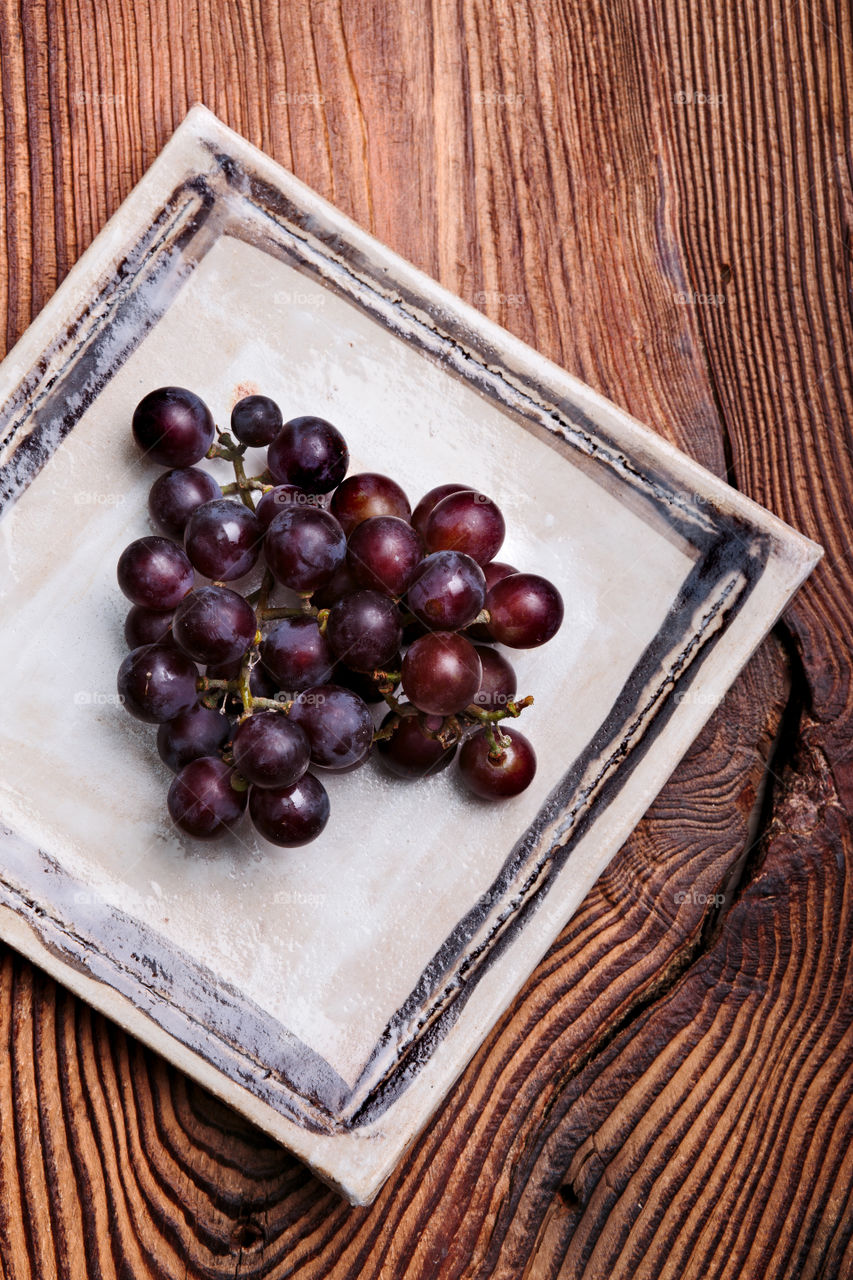Black grapes on handmade square pottery plate on old wooden table from above