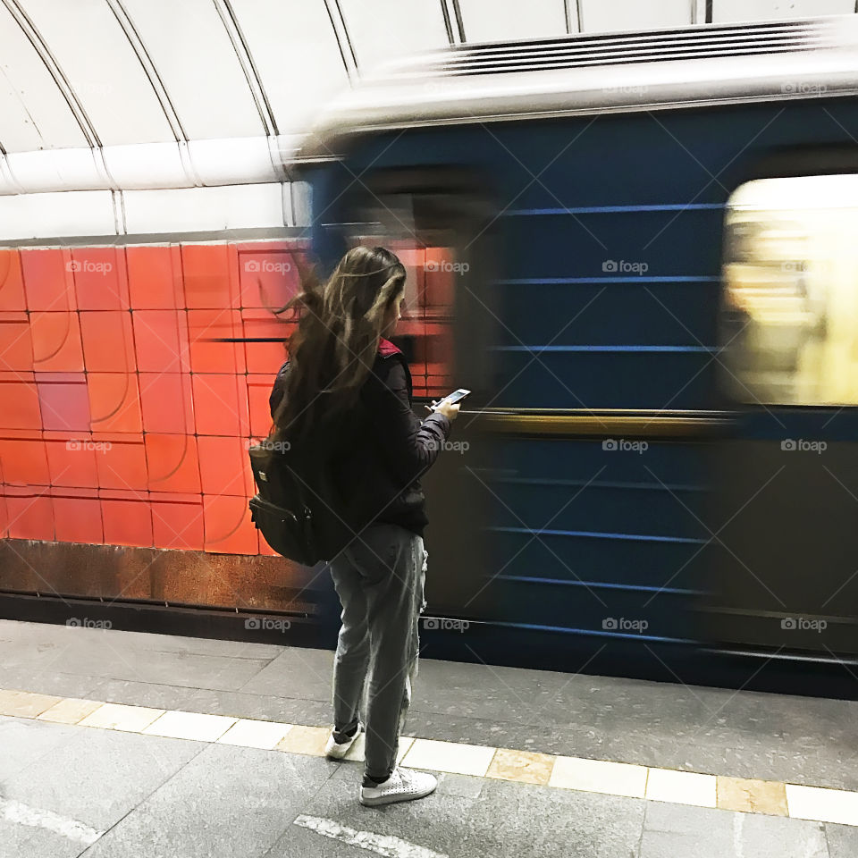 Young woman using her mobile phone in front of moving train at the subway station in the city 