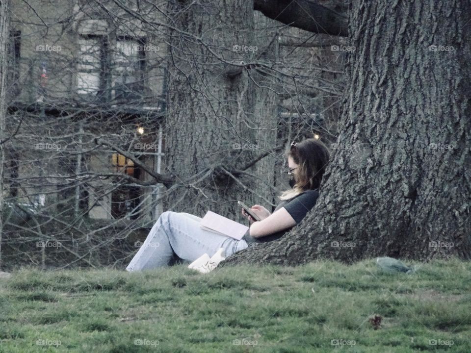 A woman leaning on the tree trunk holding a phone and a book on her lap.