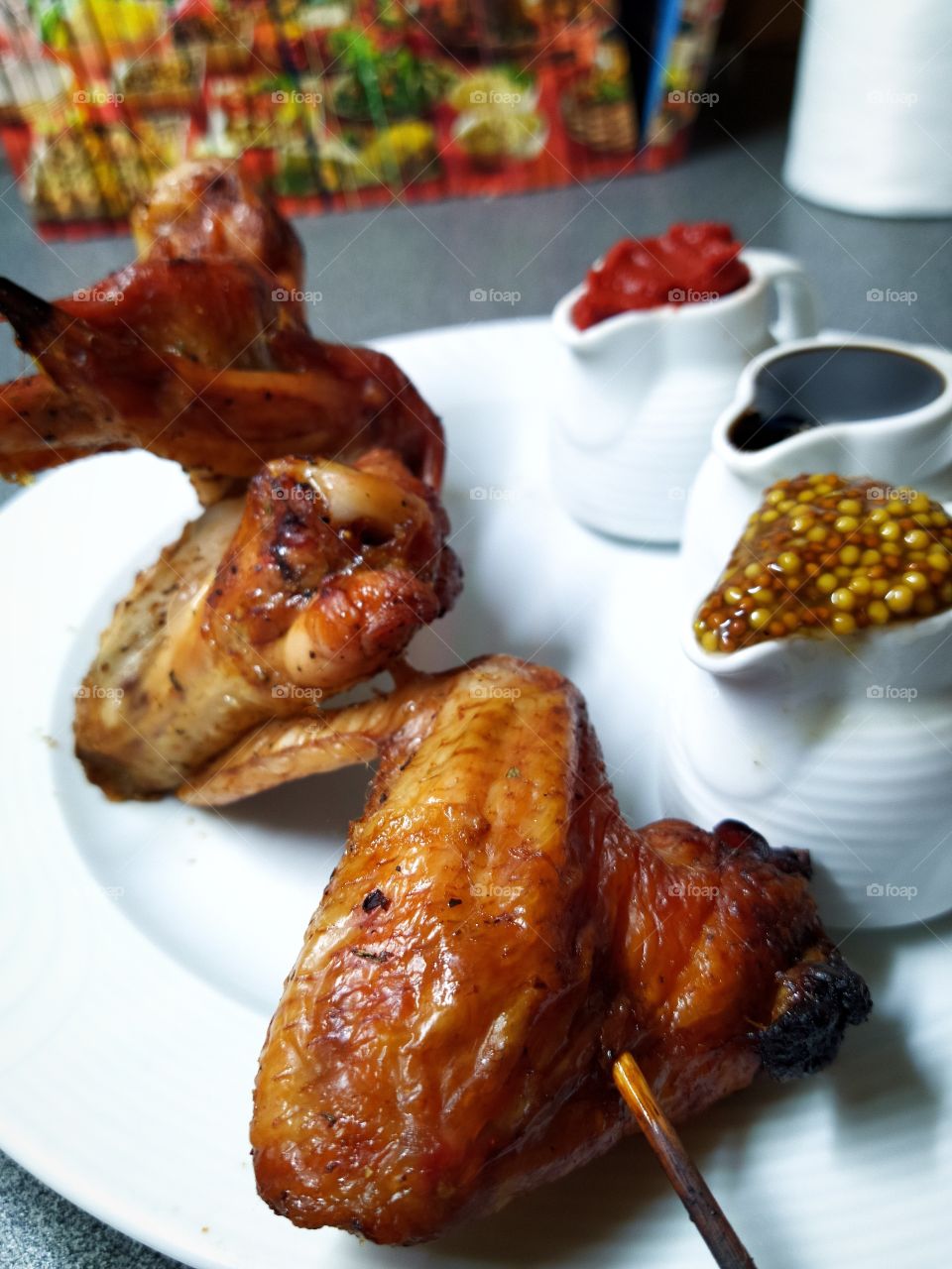 Fried chicken wings on a skewer that lie on a white plate.  Three types of sauce: French mustard, pomegranate juice and tomato sauce