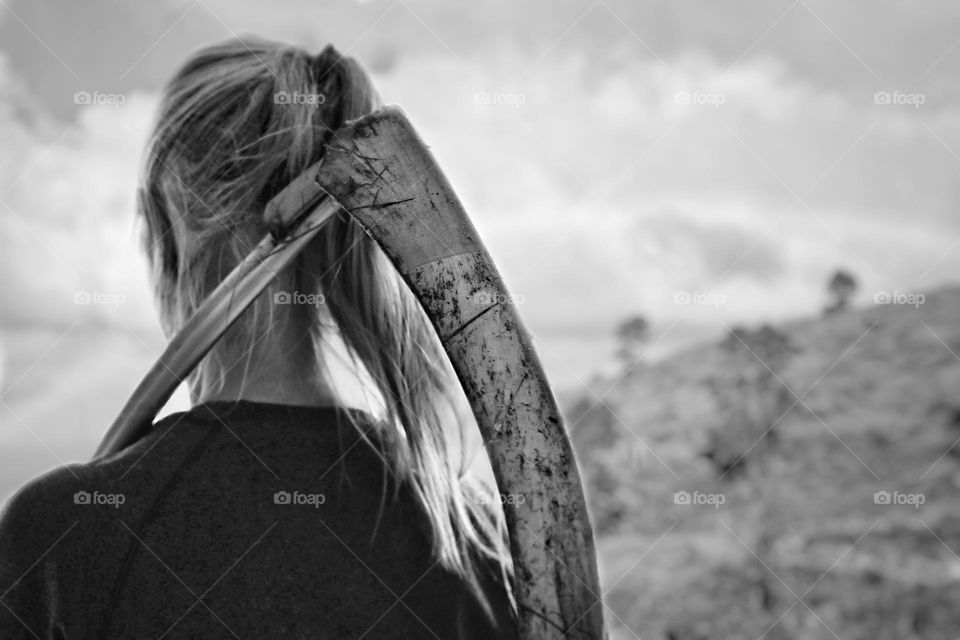 Black and white photo of a woman with a ponytail standing on a hill and carrying a scythe on her shoulder