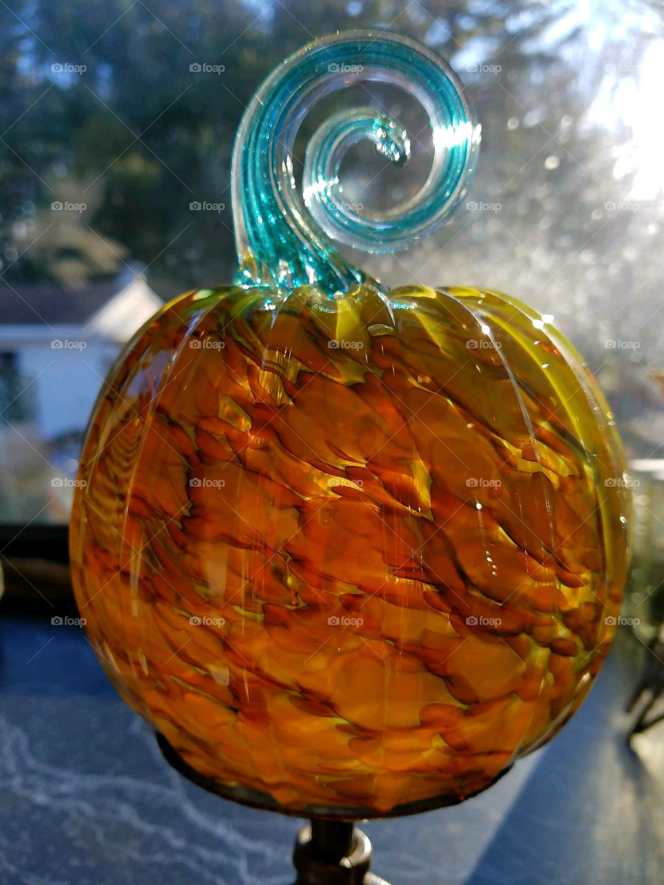 Hand blown glass pumpkin displayed on stand in sunny window. Stem is blue spiral & pumpkin is shaded with colors of orange.