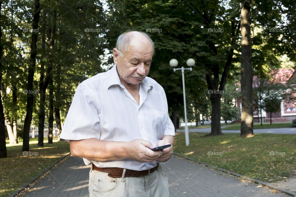 An elderly man walks alone in the park in the summer. A modern pensioner, businessman in a white shirt and trousers takes pictures with a camera in a mobile phone.