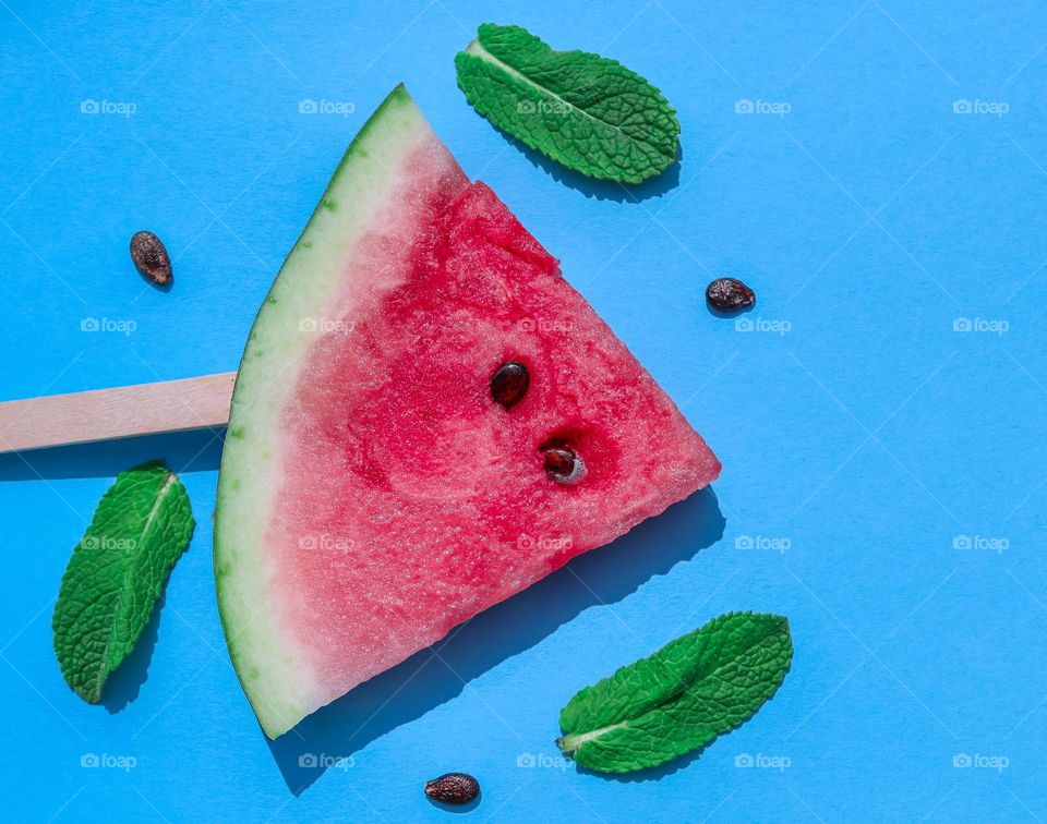 Один аппетитный спелый кусочек арбуза треугольной формы с деревянной палочкой и три листья мяты лежат на синем фоне, флат лэй копия пространство. Концепт летние угощения, фруктовое мороженое.