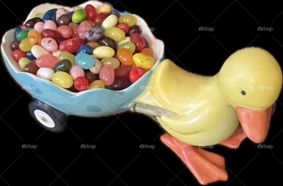A glossy yellow ceramic duckling pulls a cracked eggshell cart on tiny wheels, brimming with colorful jelly beans in red, green, yellow, and purple—sweet, playful, and perfect for springtime charm.