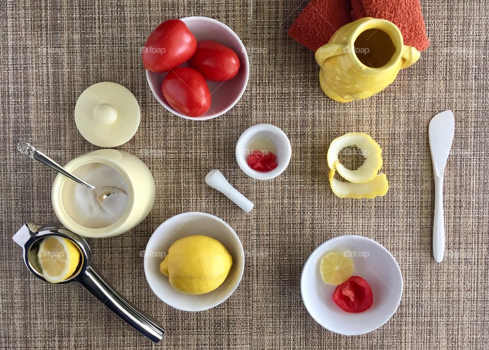 DIY Facial Mask - tomatoes, sugar and lemon in small white bowls as ingredients for a DIY exfoliating face mask with lemon peel, white mortar and pestle, orange wash cloth, white spreader, yellow vase, yellow sugar bowl, sugar spoon and citrus squeezer on natural-colored woven mat