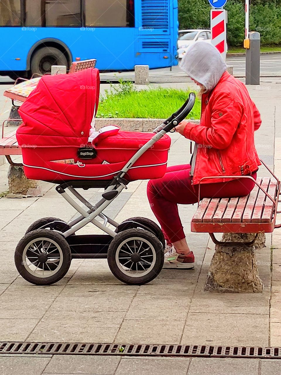 Red color.  A woman in a red jacket and red trousers sits on the Red bench.  There is a red baby carriage nearby