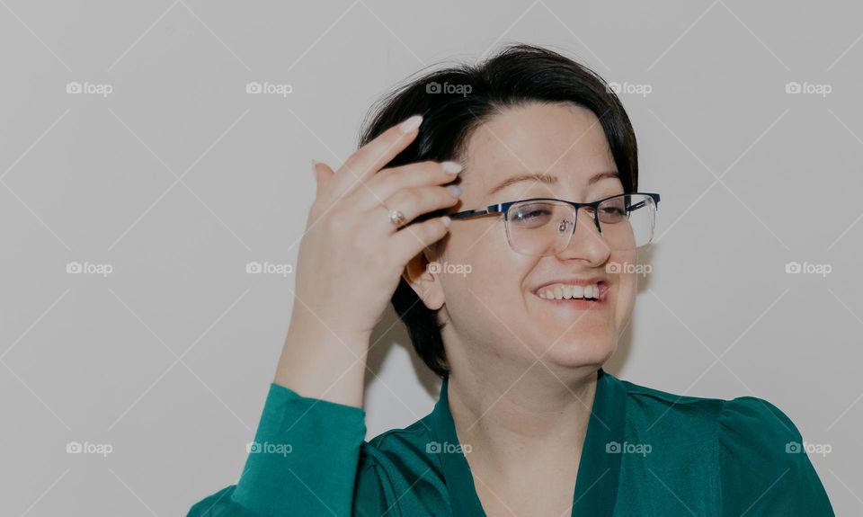 A portrait of one beautiful Caucasian brunette girl with glasses with a short haircut in a green blouse laughs happily, looks to the side and straightens her hair with her hand on a white background, close-up side view.