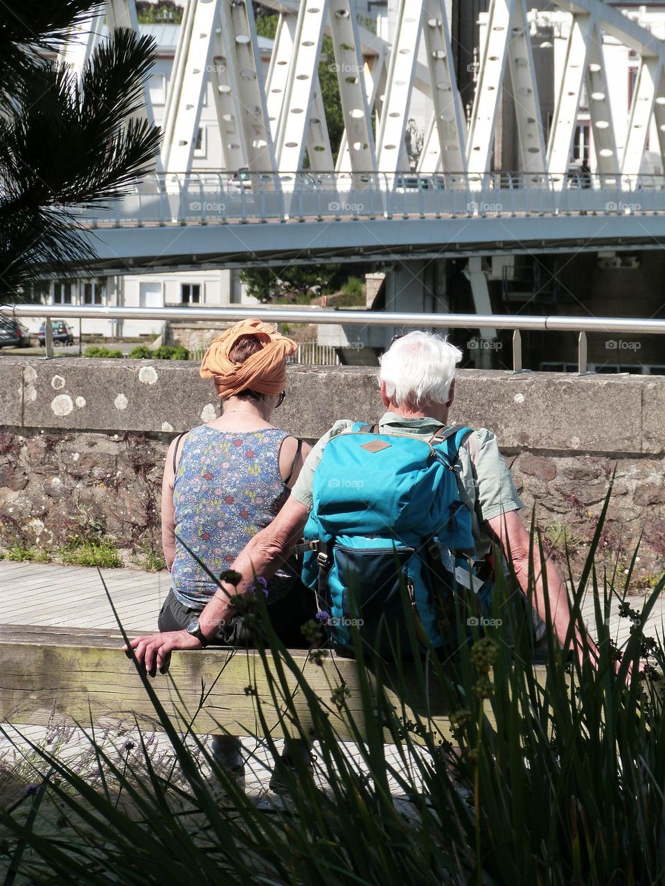 Two tourists, a senior couple, are sightseeing in town, the man is carrying a hiking backpack on his back. They are seated on a bench facing a bridge.