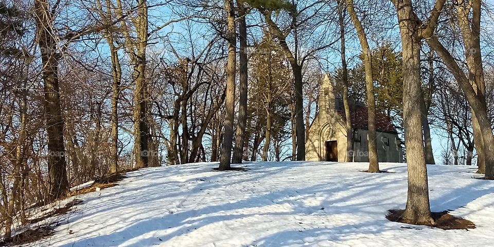 an old church in a grove of trees on a snowy hill covered in the light of the setting sun