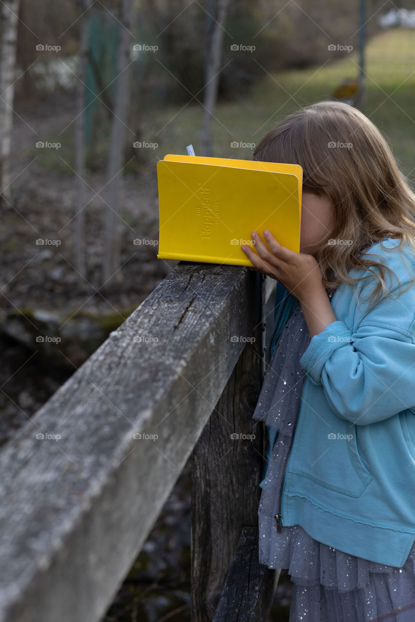Girl of 6 years is learning her HappySelf journal on a wooden bridge in the village she lives.
