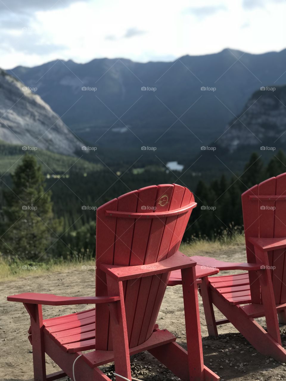 Two red wooden chairs side by side on a plateau looking at the Canadian Rocky Mountains.  Bow River can be seen running though the valley.