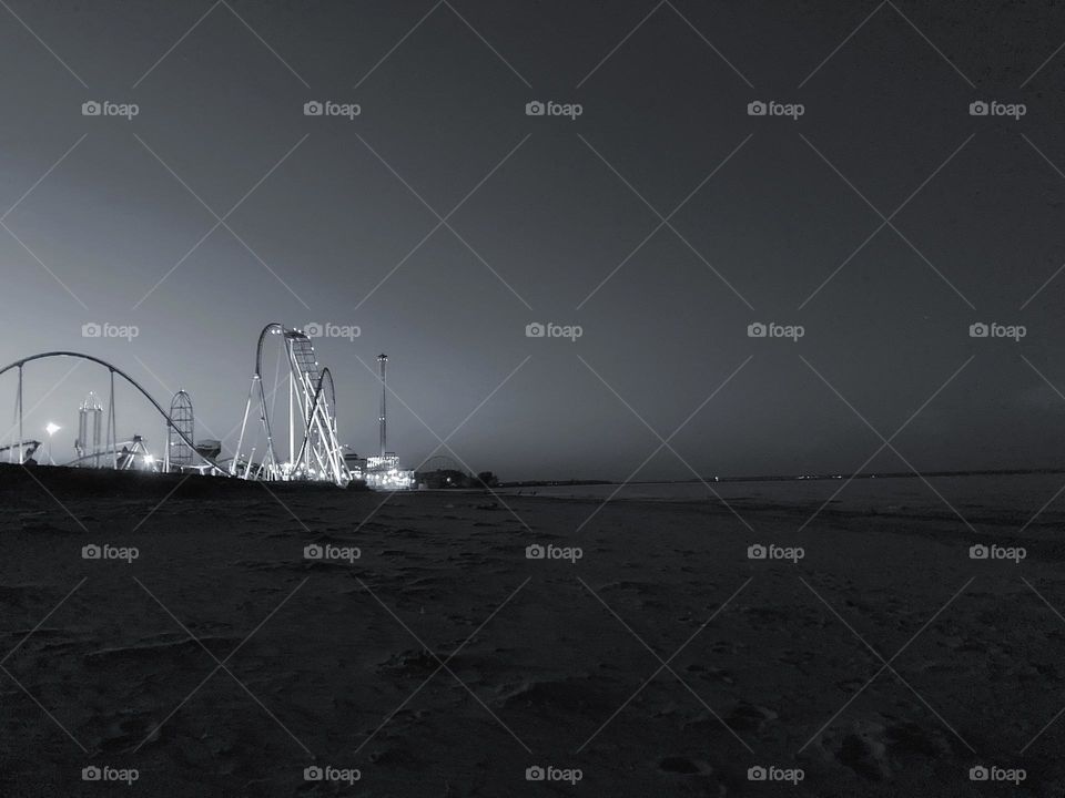 Black and white roller coasters glowing at night from sandy beach next to lake under dark sky at Cedar Point on Lake Eerie