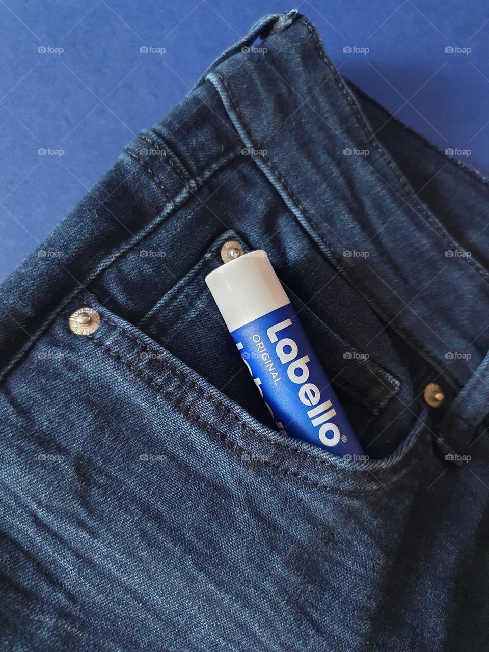 Flatlay on navy blue paper of Labello Original coming out of the pocket of a navy blue jean