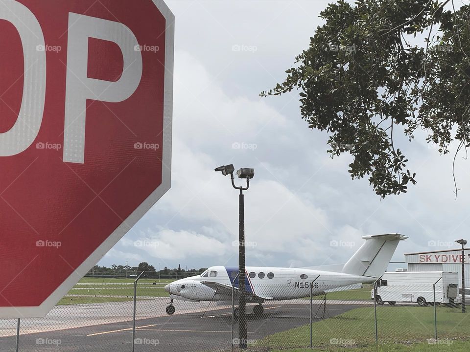 Small Airport for leisure and activities and other uses and purposes for outdoor fun and skydiving. Seeing a plane by the plane track behind a stop sign and tree.