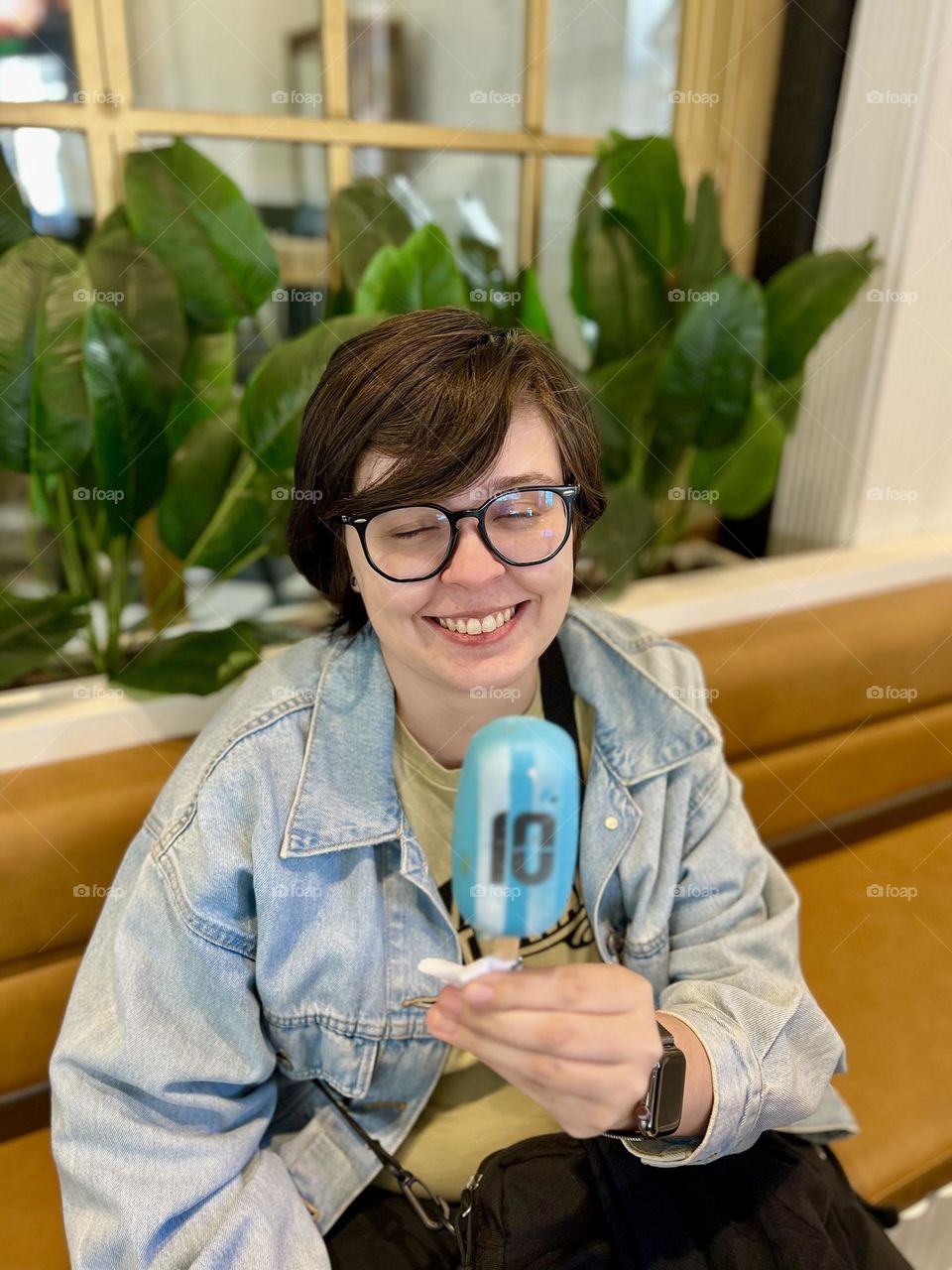 A smiling young woman holds a blue popsicle in a cozy environment. With short hair and glasses, she displays joy while sitting on a brown leather sofa, surrounded by green plants and a wall with golden frames in the background.