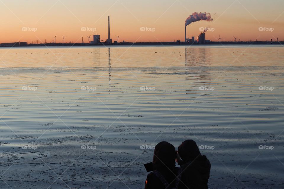 A couple stands close together in the wintry orange sunset on the North Sea with a view of the smoking chimneys of a city