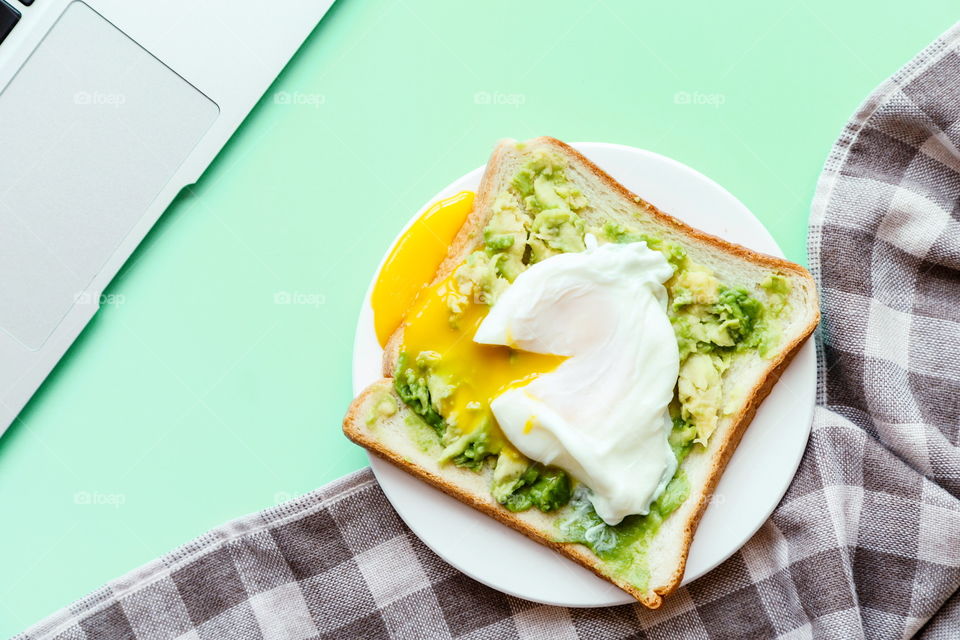 Sandwich with fresh green avocado, poached egg on white plate lying on green background 