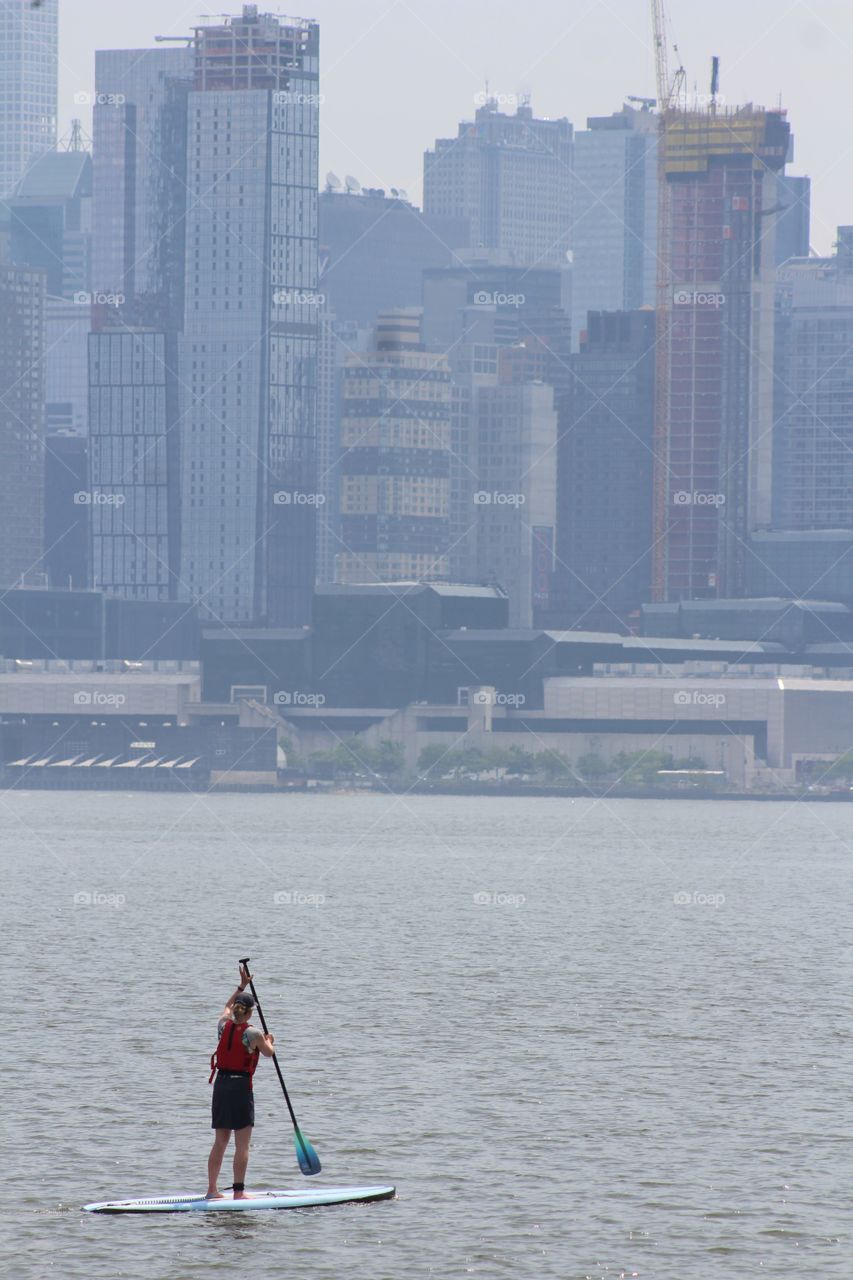 Female paddle boarder in baseball cap and life vest on Hudson River in June with New York City skyline in background 