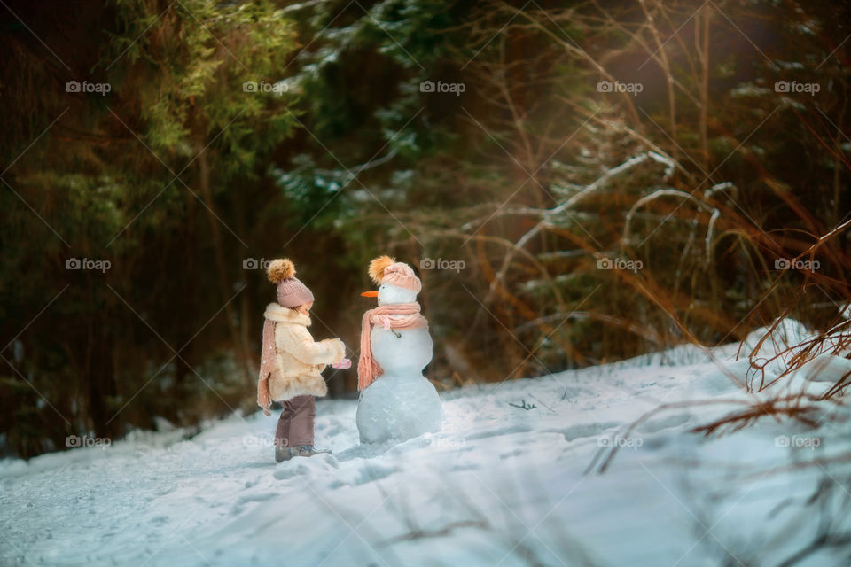 Little girl with snowman in winter forest at sunny day