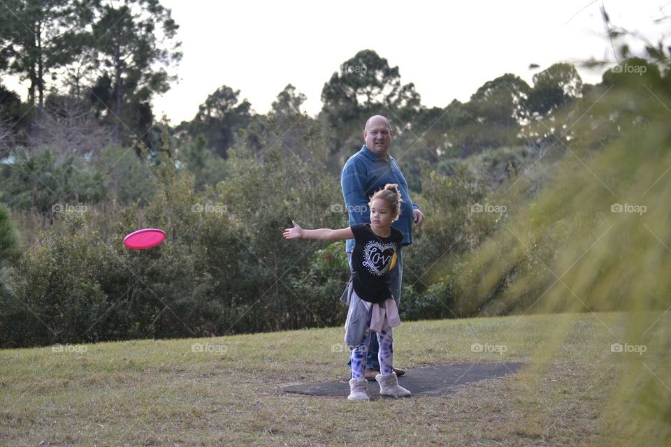 A little girl throwing a pink frisbee with her grandfather standing behind her at a park
