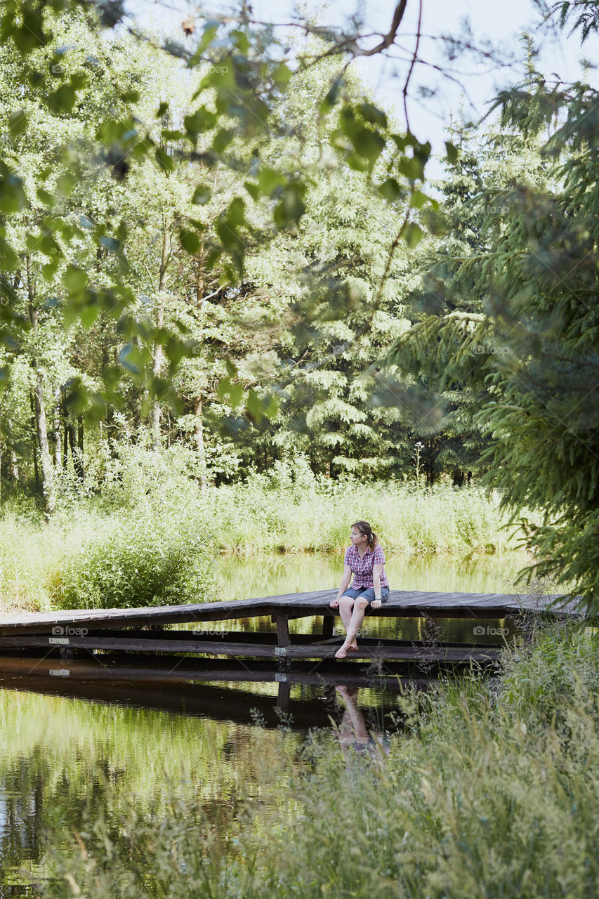 Woman sitting on a bridge over a lake, among the trees, close to nature, during summer vacations. Candid people, real moments, authentic situations