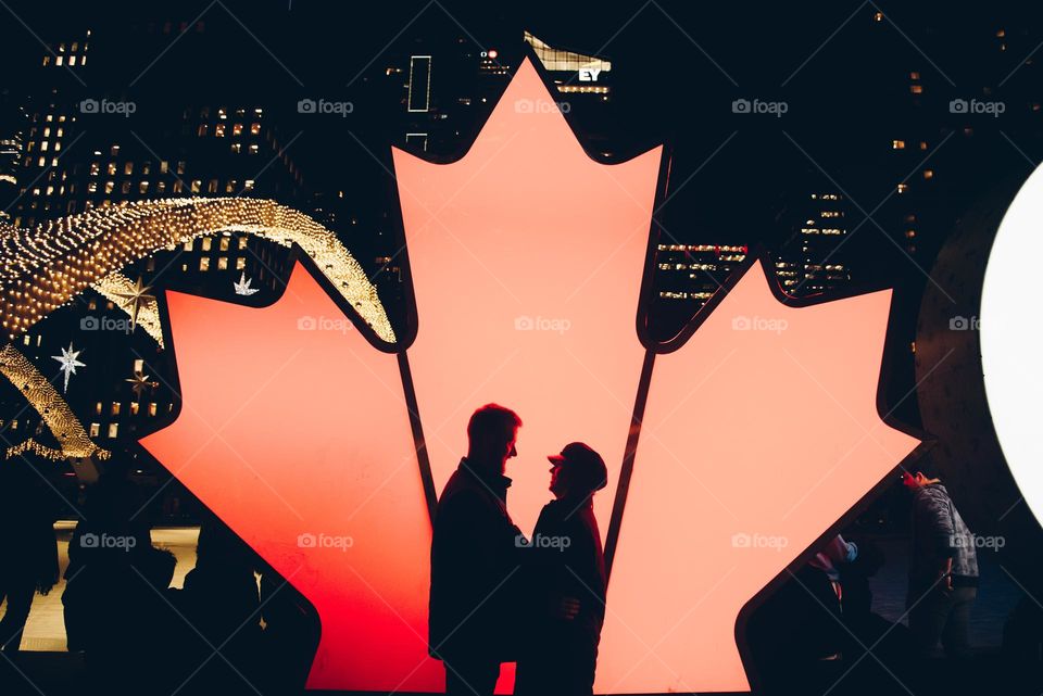 glowing hearts; two people in front of Canadian Maple leaf at Nathan Philips square with Toronto skyline in background and light glowing on face
